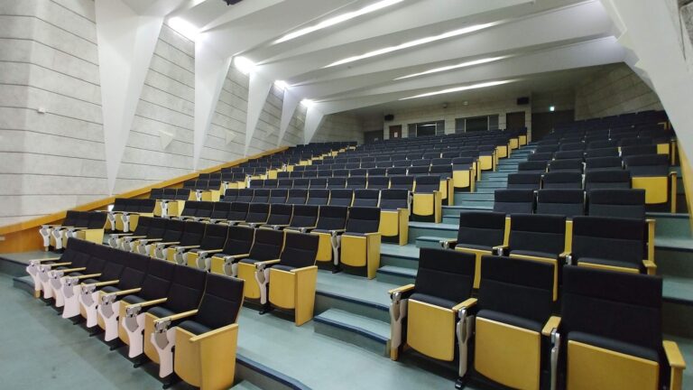 A tiered university lecture hall viewed from the back row with half-full seats, a speaker at the distant lectern, and open laptops glowing on desks.