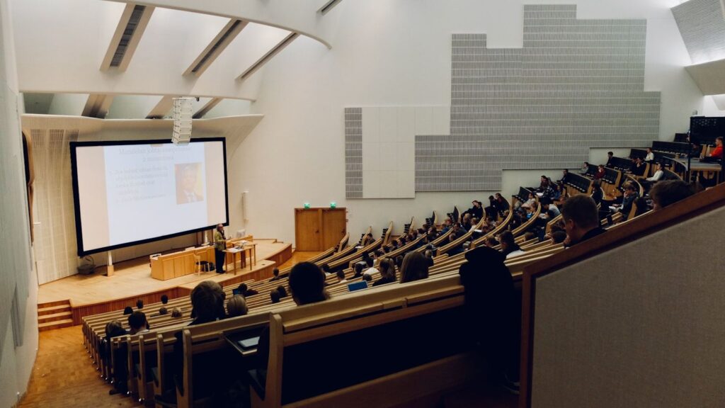 An empty university seminar room with a large monitor showing a paused recorded lecture and a single notebook open on the table.