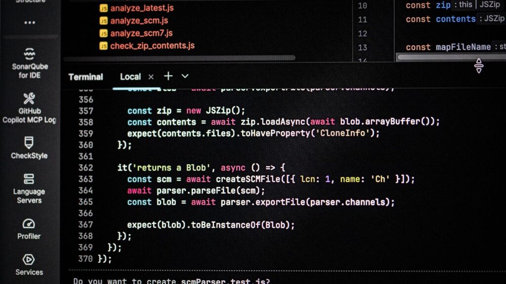 A laptop open to a code editor on a small desk beside a mug, with a closed textbook and a printed schedule taped to the wall.