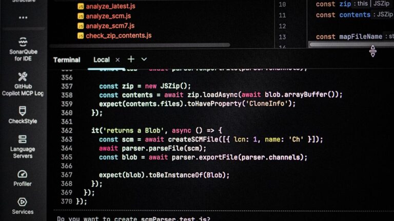 A laptop open to a code editor on a small desk beside a mug, with a closed textbook and a printed schedule taped to the wall.
