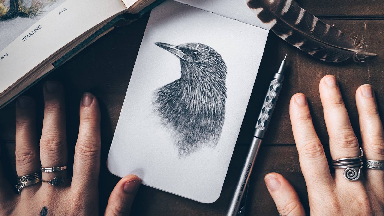 An open sketchbook on a wooden desk beside a pencil and an open textbook, showing a rough process diagram drawn in pencil.