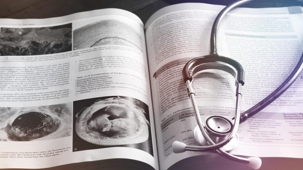 A medical student in scrubs holding a phone displaying a flashcard with a pharmacology question, leaning against a hospital corridor wall.