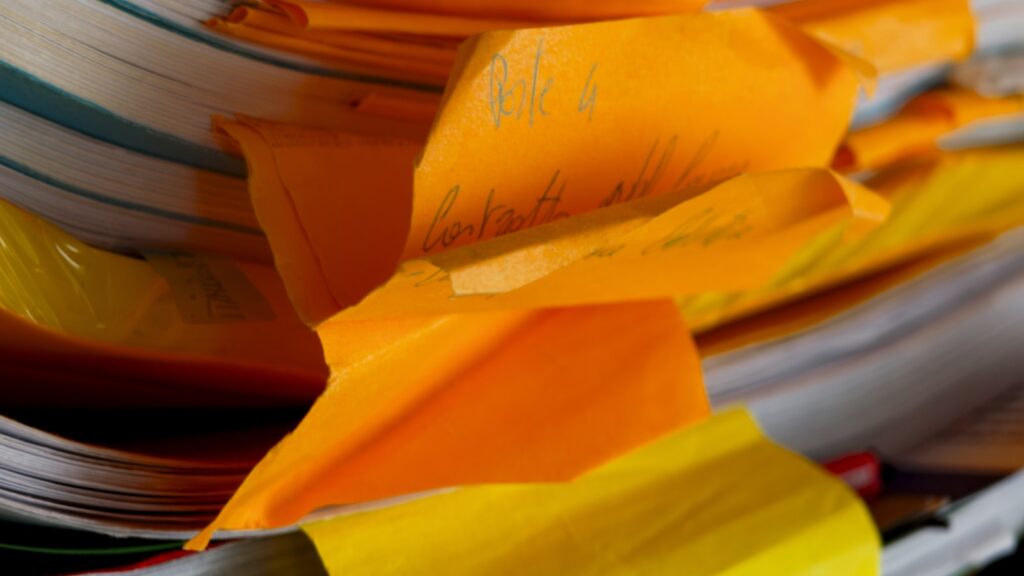A desk cluttered with printed journal articles, sticky notes in three colors, and a laptop open to a reference manager with hundreds of entries visible.