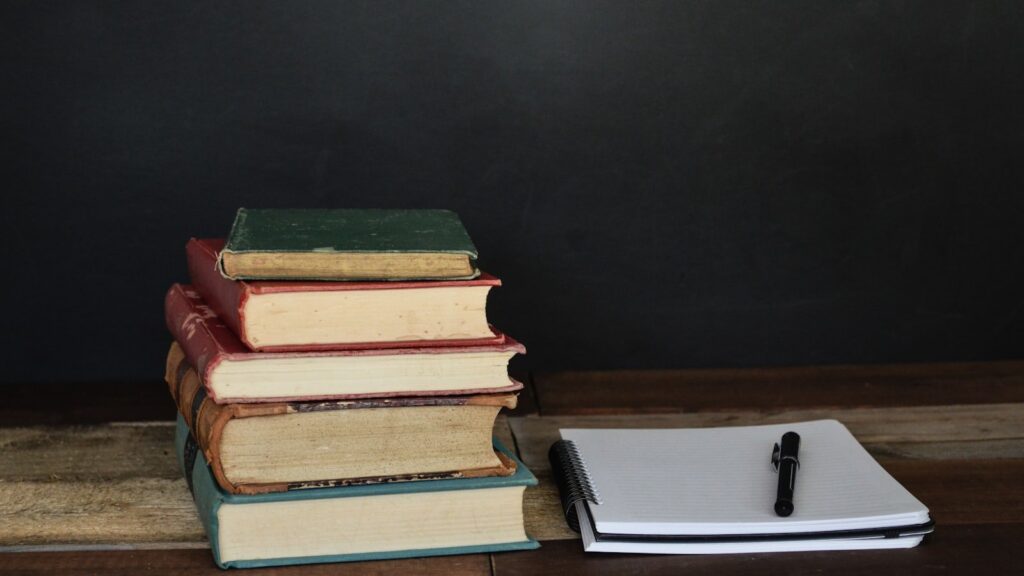 An open library book rests beside a laptop screen showing dense reference text in a quiet reading room.