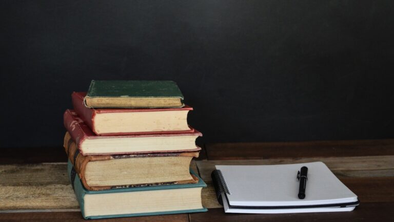 An open library book rests beside a laptop screen showing dense reference text in a quiet reading room.